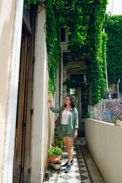 vertical image of young woman standing in entrance of her house caressing leaf of wall creeper plant