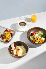 A diverse breakfast spread with millet porridge, salad bowl, syrniki, Americano coffee, and orange juice in sunlight