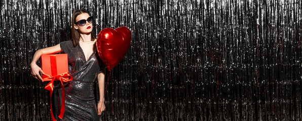 Beautiful fashionable woman in a shiny glamour little dress with a heart-shaped balloon and a gift...