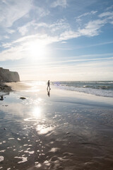 This photograph captures a solitary figure taking a peaceful walk along the shore, bathed in the ethereal glow of the coastal sunlight. The sun, hanging low in the sky, casts a brilliant reflection on