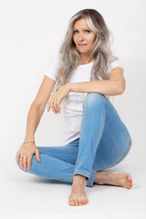 A beautiful elderly woman with gray long hair in jeans poses while sitting on a white background.