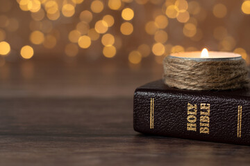 Candle burning and closed holy bible book on wooden table with bokeh background. Close-up. Copy...