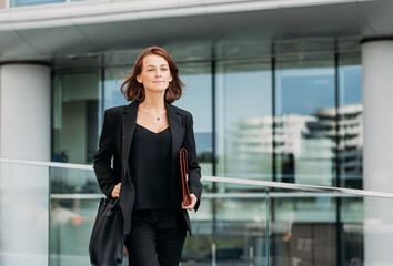 Fototapeta premium Confident female with a bag and leather clothes walking outdoors against a glass building. Cheerful corporate person walking outdoors.