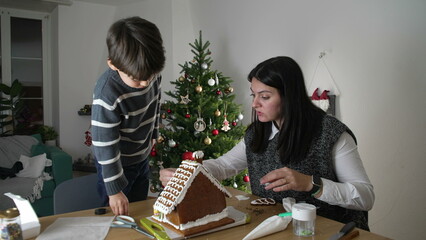 Mother and Child Creating Gingerbread House, Home December Festivities with Christmas Tree in background. Family preparing for holidays