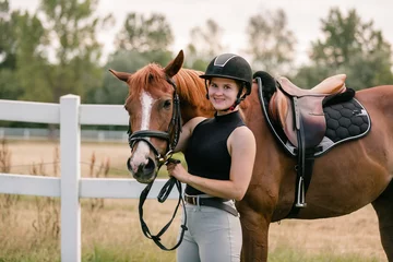 Foto op Canvas Female rider in equestrian clothes holding the reins and leading her beautiful saddled chestnut horse. Horseback riding activity concept. © 24K-Production