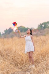Girl in White Long Dress Holding a Colorful Windmill Toy in a Dry Grass Meadow