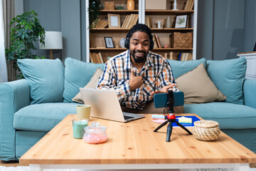 Happy millennial African American man in headset having web call using laptop computer and smartphone sitting on couch talking at webcam in home office. Video conference online business meeting.
