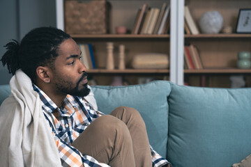 Young depressed African American man sitting on sofa with crossed arms. Racism victim for skin color black male suffering from depression after being fired from work Job lost and mental health concept - obrazy, fototapety, plakaty