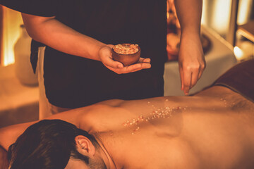 Man customer having exfoliation treatment in luxury spa salon with warmth candle light ambient. Salt scrub beauty treatment in Health spa body scrub. Quiescent