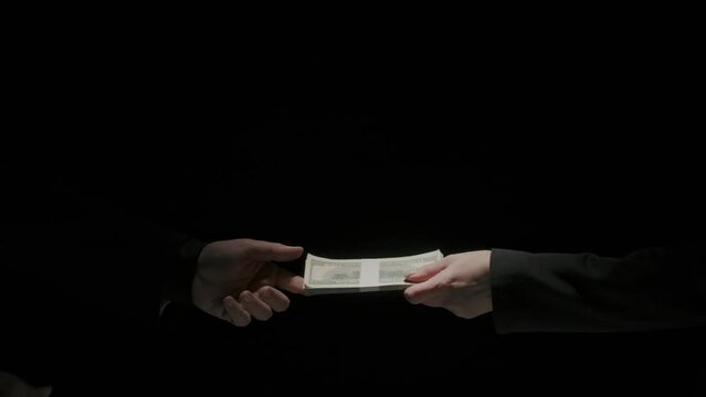 Portrait of business people in suits on black background. Partner gives to female colleague wad of dollar banknotes making deal handshake.