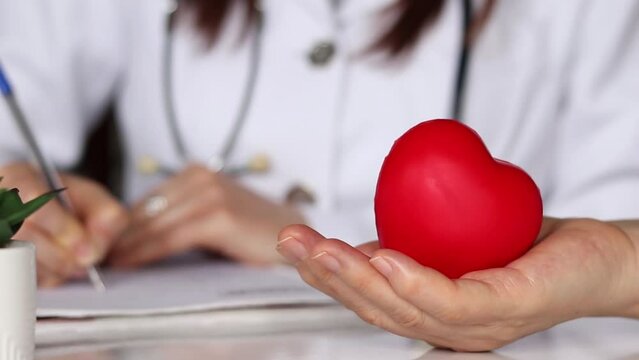 Patient holding red heart in hand while a woman cardiologist doctor is writing a medical diagnosis report in the background. Close-up. National Cardiology Day concept.