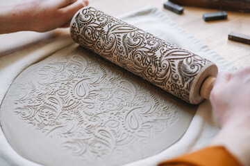 Potter's hands embossing a pattern on a clay plate with a rolling pin Decorating a clay blank with a ornament