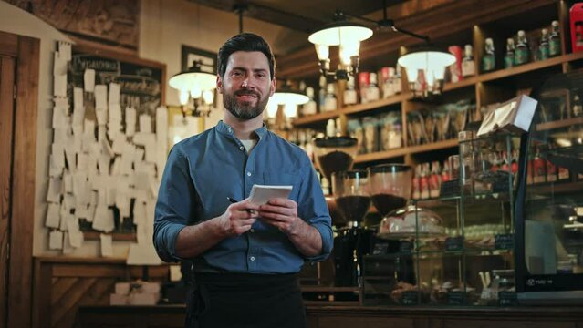 Portrait of friendly bearded man in uniform writing down order on paper notes while standing at stylish vintage bar. Competent male waiter smiling and looking at camera during working process.