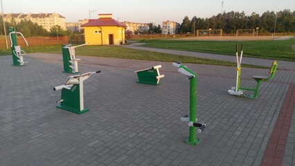 Obraz premium A variety of metal exercise equipment for general use is set up on a concrete paved area next to a lawn, sandy football field, metal fence, buildings and trees. The summer sun is shining at sunset