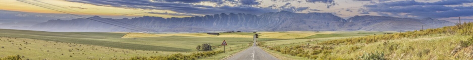 Panoramic image of a paved, empty road running straight towards a mountain range on the horizon