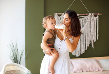 Young loving mother holding child hands and feeding baby boy son with milk from bottle at home
