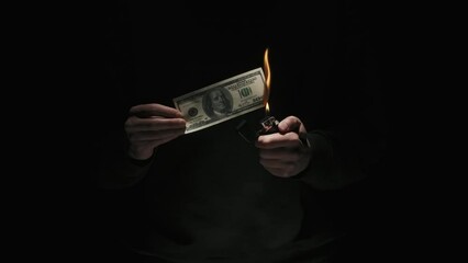 Portrait of male in black clothes isolated on black background in low light. Man holding hundred dollar banknote lights up lighter and burns money.