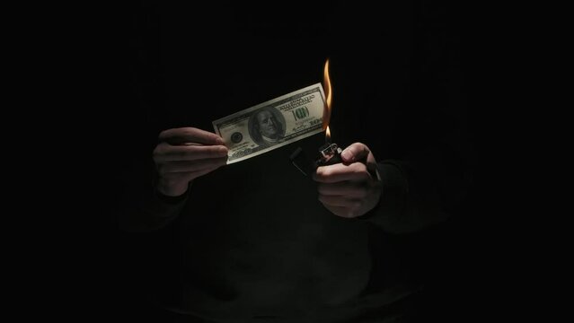 Portrait of male in black clothes isolated on black background in low light. Man holding hundred dollar banknote lights up lighter and burns money.