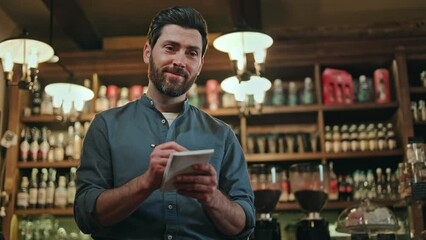 Attractive caucasian man dressed in blue uniform smiling and talking pleasantly while taking guest order at stylish cafe. Competent male waiter with beard recording details on paper notepad using pen.