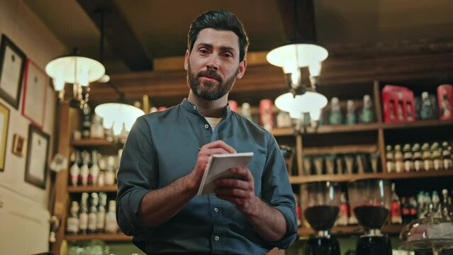 Charismatic male waiter in blue shirt and apron using pen and notes while servicing customers at retro style bar. Caucasian dark haired man giving professional advice about menu and repeating order.