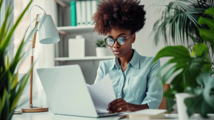 Young woman focused on her work at a desk with laptop and holding documents - obrazy, fototapety, plakaty