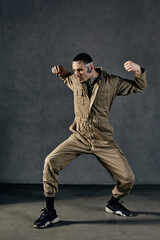 Active male with tattooed body and face, earrings, beard. Dressed in khaki jumpsuit and black sneakers. Dancing on gray background. Dancehall, hip-hop