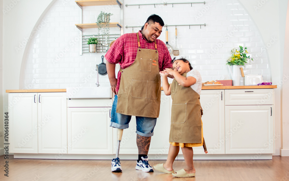 Wall mural Portrait Asian two people of father with handicap prosthetic leg, little daughter playing, standing in kitchen at cozy home, looking each other, smiling with happiness, love. Family Concept.