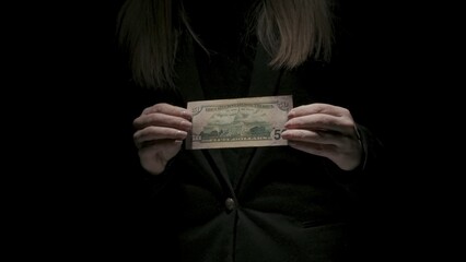 Portrait of female in black suit isolated on black background in low light. Woman holding in hands fifty dollars banknote backside at the camera.