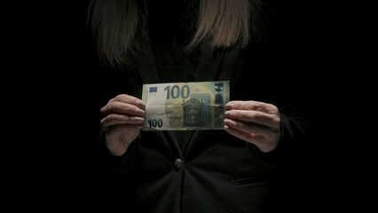 Portrait of female in black suit isolated on black background in low light. Woman holding stretching in hands hundred euro banknote at the camera.