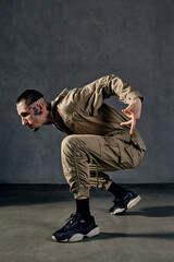 Attractive performer with tattooed body, earrings, beard. Dressed in khaki overalls and black sneakers. Dancing on gray background. Dancehall, hip-hop