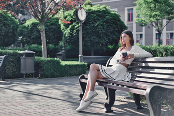 Time for contemplation and decision-making. A beautiful young adult lady holds a smartphone in her hands and thoughtfully looks to the side while sitting on a bench in a quiet city park