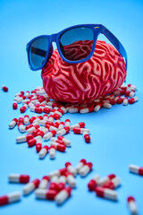 Red brain with sunglasses on and a bunch of pills around it, on top of a blue surface.