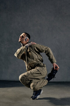 Extraordinary guy with tattooed body, earrings, beard. Dressed in khaki overalls and black sneakers. Dancing on gray background. Dancehall, hip-hop