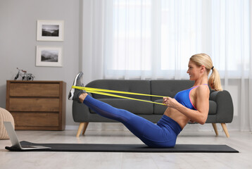 Fit woman doing exercise with fitness elastic band near laptop on mat at home