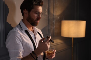Handsome man with glass of whiskey smoking cigar at home. Space for text