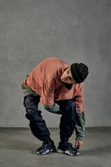Athletic man with tattooed body, earrings, beard. Dressed in hat, casual clothes and black sneakers. Dancing on gray background. Dancehall, hip-hop