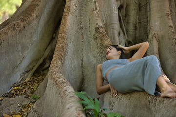 South American woman, young, pretty, brunette in gray top and skirt lying sleeping on the roots of a big tree, relaxed and calm. Concept beauty, fashion, diversity, peace, nature.