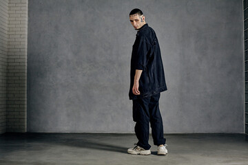 Obraz premium Athletic fellow, tattooed body, beard. Dressed in casual clothes, white sneakers. Looking back, posing on gray background. Dancehall, hip-hop
