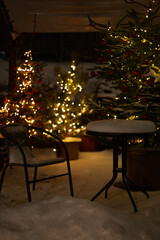 Snowy patio with Christmas trees, lights, chair and table at night. Christmas card