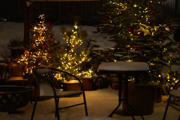 Beautiful winter view with Christmas trees, snow, lights, chair and table on the patio at winter night. Vintage