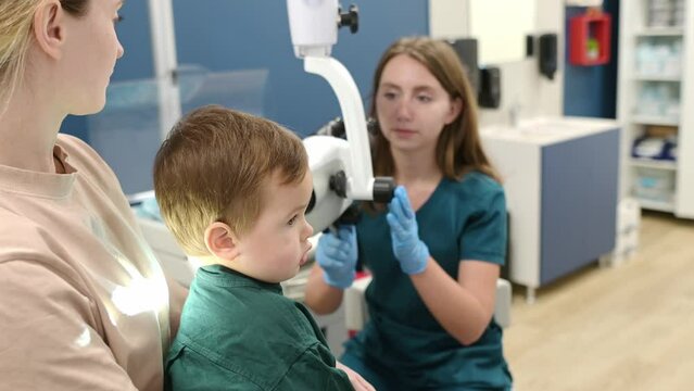 A cute toddler boy with his mother are at an appointment with a pediatric ENT to prepare for preschool. The doctor otolaryngologist is examining the ear of a little patient under ENT microscope