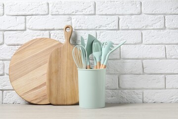 Different cutting boards and kitchen utensils in holder on light table near white brick wall. Space for text