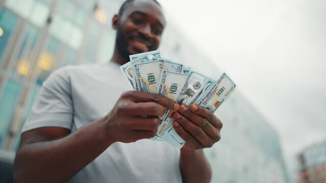 Man counts dollars banknotes on street. Contented smiling african american guy enjoying earned cash money in city business center, low angle view. Finances, wages concept. Entrepreneur, businessman.