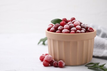 Frozen red cranberries in bowl and green leaves on white table, closeup. Space for text