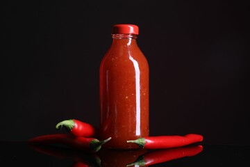 Spicy chili sauce in bottle and peppers against dark background