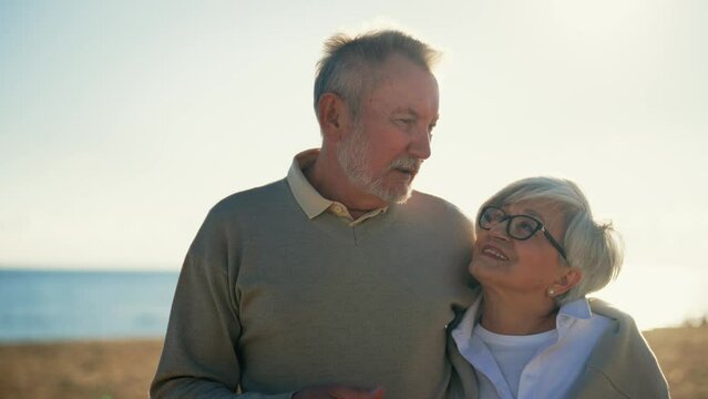 Old senior happy smiling man woman looking at camera. Husband tenderly hugging wife. Portrait elderly positive couple rests at sea in sunny day. Love romantic relationships lifelong marriage concept.