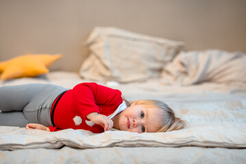 Cute baby toddler girl child in a red suit lies on the bed during the Christmas holiday at home.
