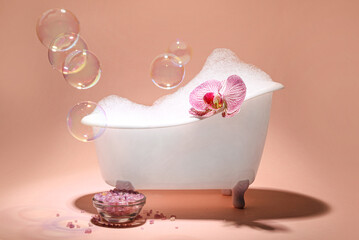 Small bathtub with foam, orchid flower, soap bubbles and sea salt on nude background
