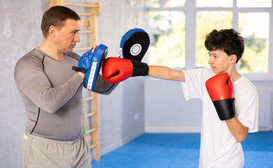 Sportive teenage boy training boxing kicks on punch mitts held by instructor in sports hall