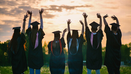 Silhouettes of graduates in black robes waving their arms against the evening sunset.